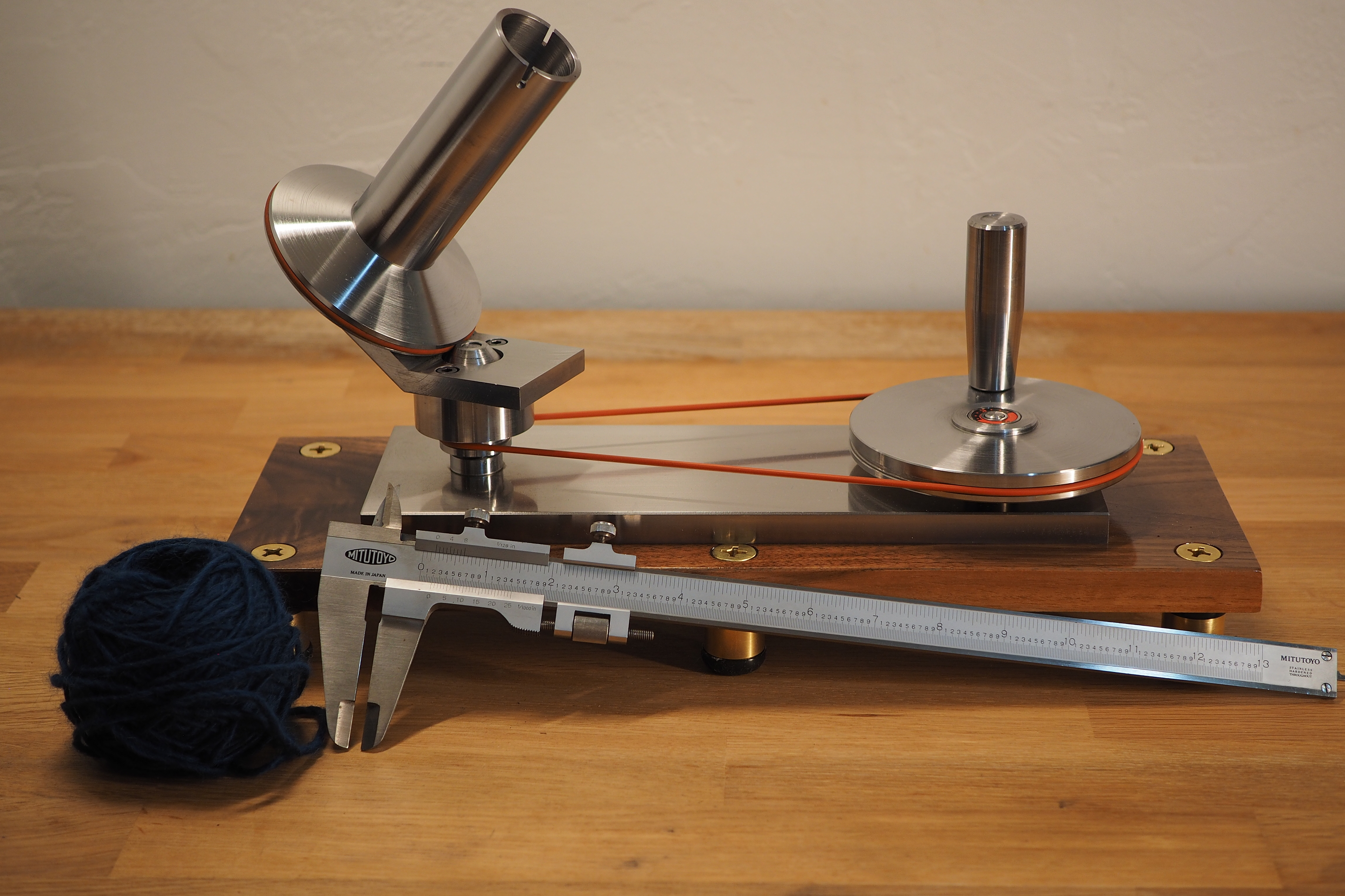 A picture of a very shiny solid steel yarn winder on a wooden surface. A ball of yarn and some vernier calipers are in the foreground.