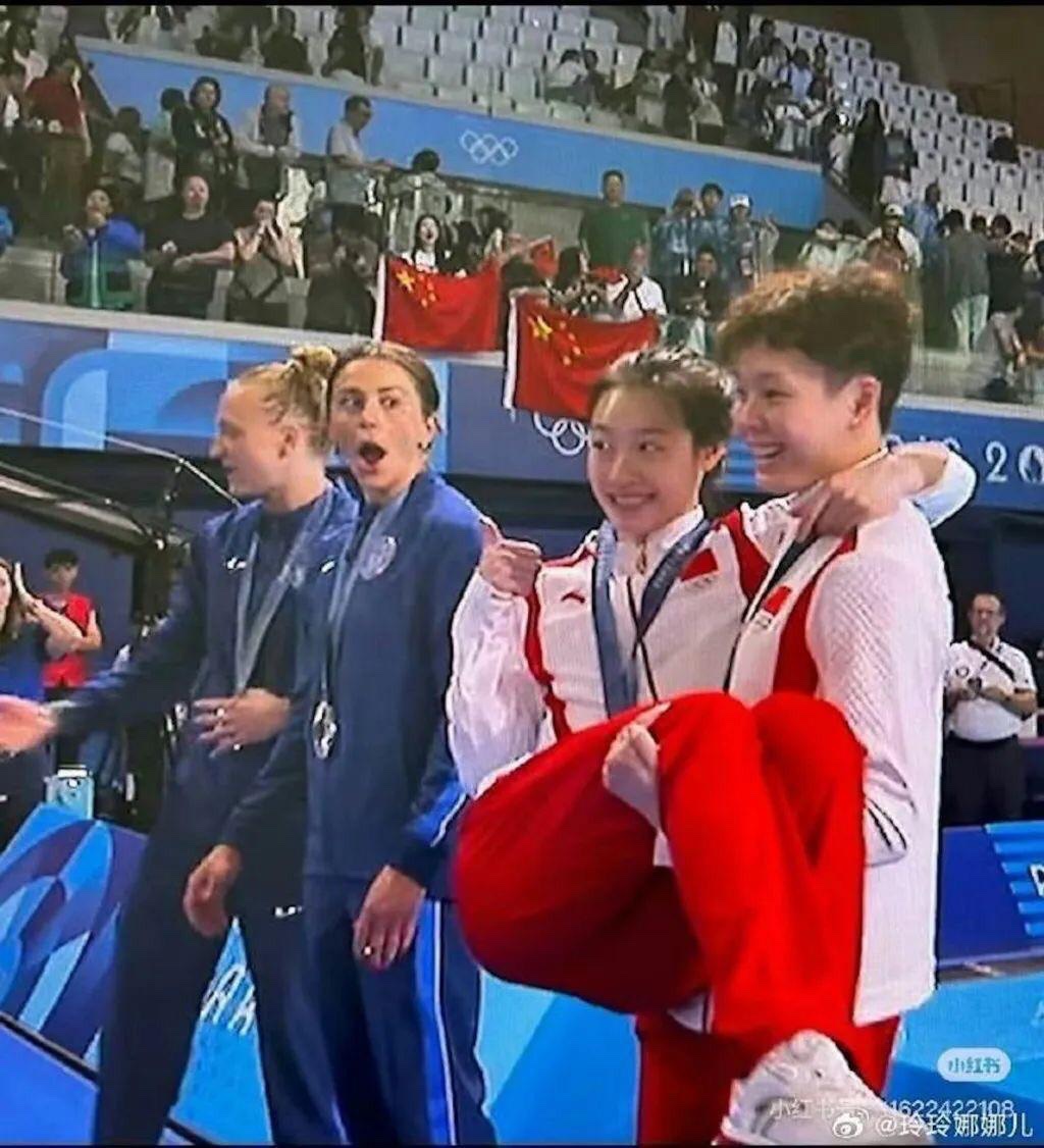 At a podium of the Olympics women’s synchronized springboard (paired), a US athlete is amazed while looking at a Chinese athlete scooped up at the legs by another Chinese athlete