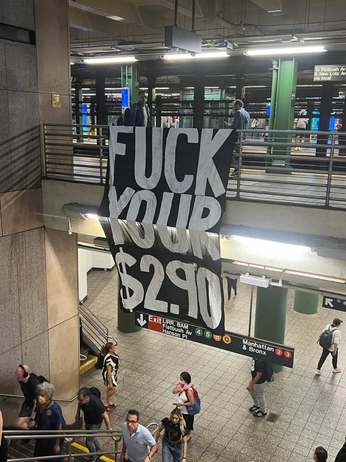 photo taken inside an NYC subway station. there is a huge banner hung from an upstairs railing that reads "FUCK YOUR $2.90"